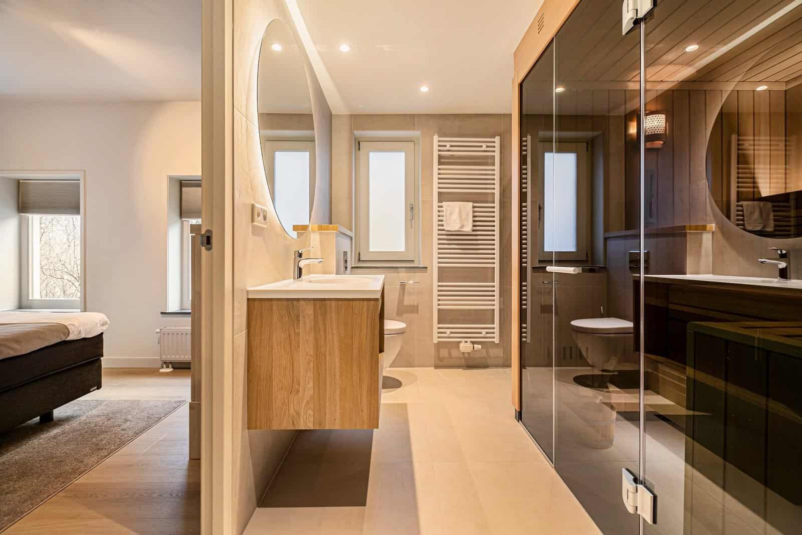 Zwevend eiken badmeubel met wit solid surface wastafelblad en opstaande waterkering tegen de wand, naast een sauna met glaswand, Hof van Saksen